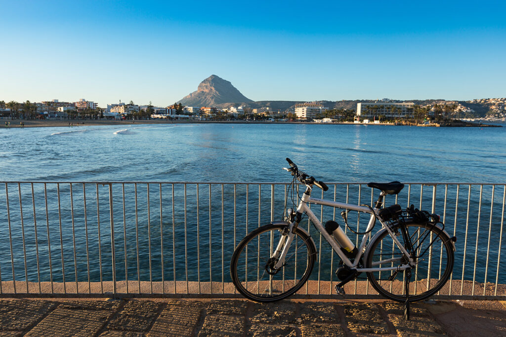 Fahrrad an einer Brüstung mit Meer und Bergen am Strand El Arenal in Xàbia, Region Valencia.